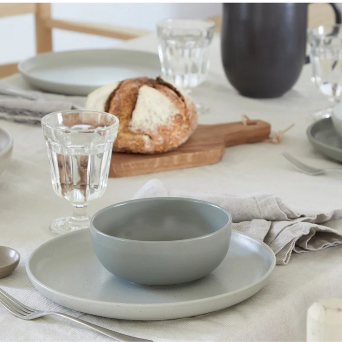 A dining table set with Costa Nova Pacifica Dinnerware in Oyster Grey—gray ceramic bowls and plates—paired with clear glasses, silverware, and a round loaf of bread on a wooden board over a light tablecloth.